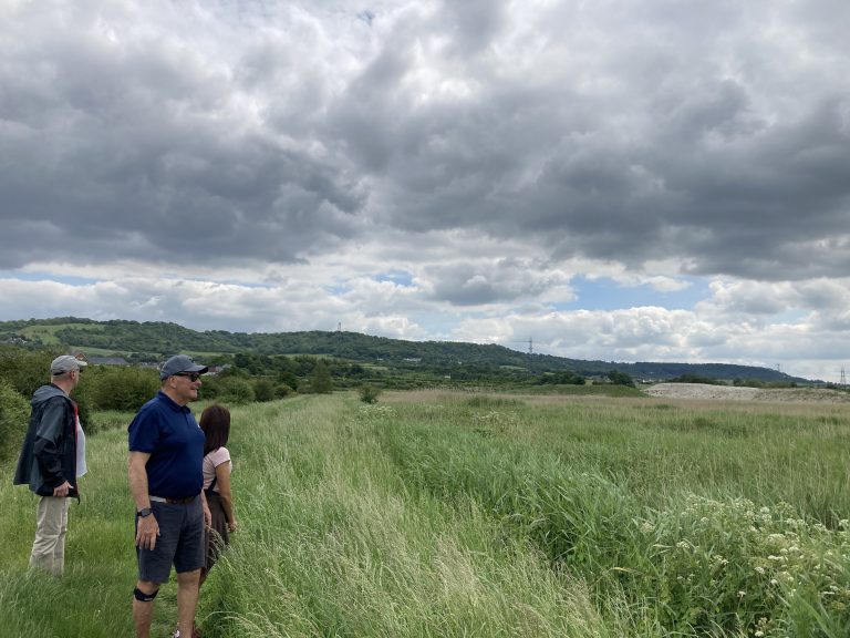 Three people on top of a grassy riverbank. To their right are reed-choked mudflats. Behind then a hedgerow follows the curve of the riverbank. Green hills rise steeply in the distance. The sky is full of light grey clouds with small pockets of blue between them.