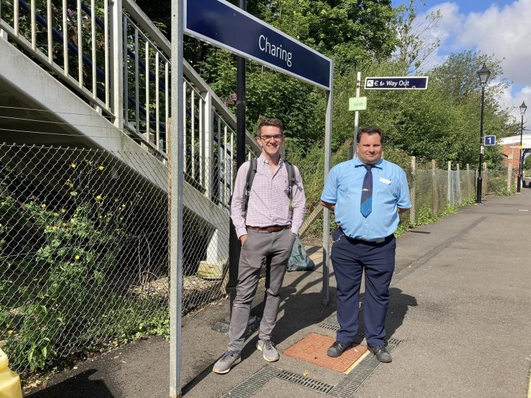 Jonny and Mark stood on the platform beneath a "Charing" station sign. Behind them is a fence line between the platform and an overgrown area of trees and shrubs.