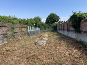 A wide strip of ground surrounded by walls and a chain-link fence. The site shows signs of recent vegetation clearance. There is a row of four ton bags containing soil that runs through the middle of the site