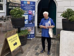 Volunteer Chair Linda Brinklow wearing a Kent Community Rail Partnership polo shirt. Linda is standing in front of a doorway next to which stands a Kent CRP promotional roller banner. A sign points to the door "Model Railway Exhibition"