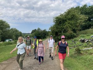 A group of walkers on a wide gravel path between grass verges. To their left are boats moored on a river