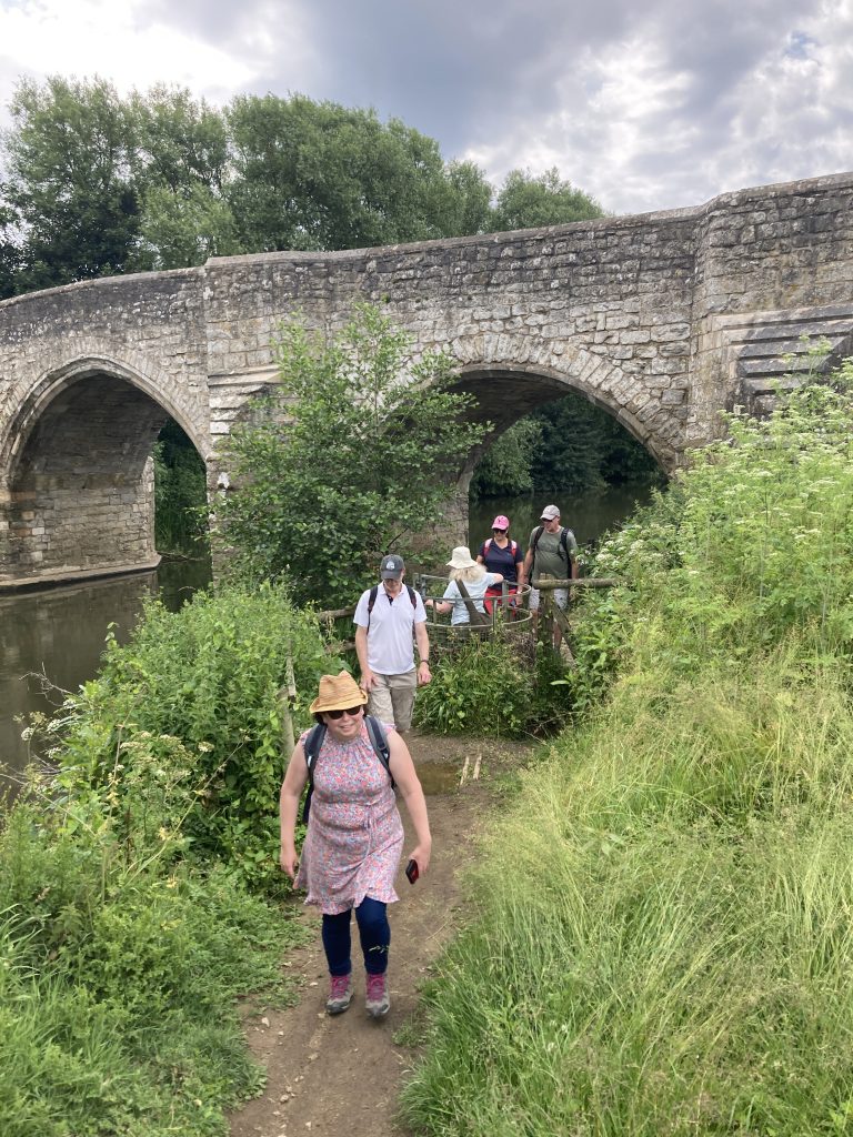 Walkers on a narrow path negotiate a kissing gate. Behind them is a stone bridge with arched spans crossing the river
