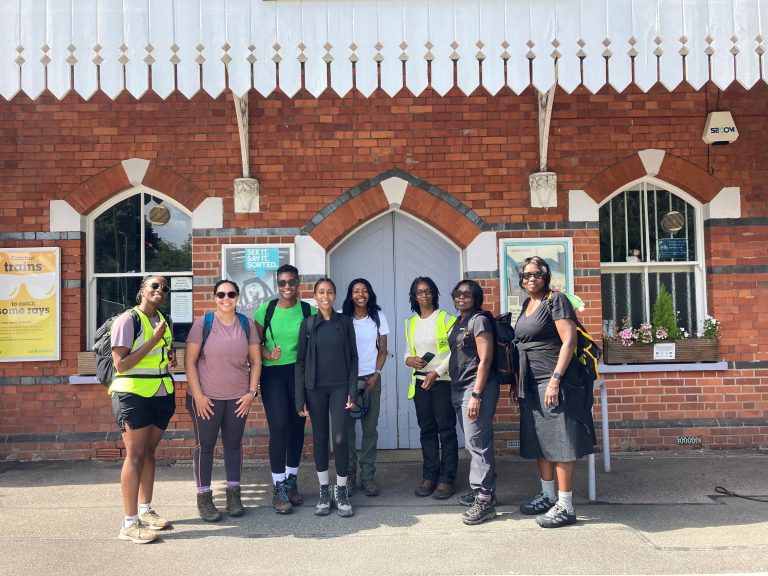 A group of people pose for a picture in the sunshine beneath a wooden canopy shelter outside a station building. There are window boxes of flowers at windows either side of the closed entrance doors.