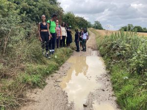 A group of walkers stand beside a large muddy puddle on a gravel trail. The sky above is thick with dark grey clouds