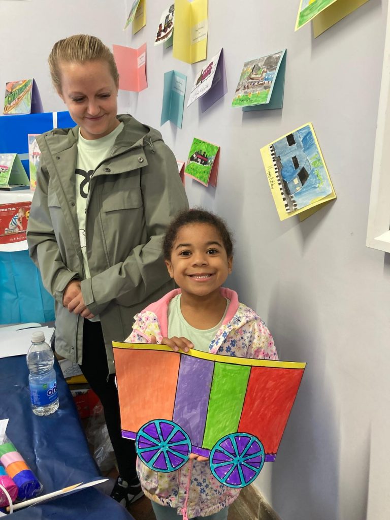 A young person holding their decorated train-carriage poses for a picture outside inside Queenborough station booking hall. A parent stands behind them. There are hand made birthday cards on the walls.