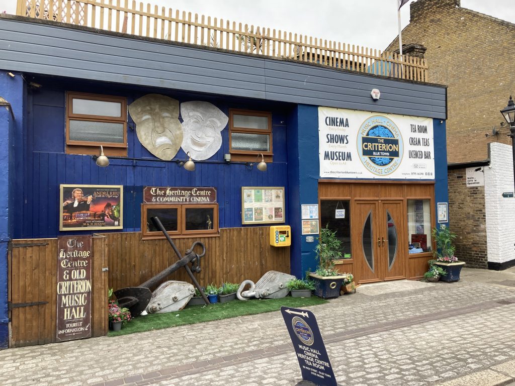 The exterior of the Criterion heritage centre and community cinema in Bluetown. A theatre building with wood cladding. The top floor is painted blue. There are large Tragedy and Comedy masks between the top floor winfows. Outside are a shps anchor and two large pulleys. Potted plants have been placed wither side of the entrance doors.