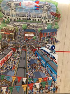Wall mural inside Inspiration: A "Where's wally" style crowd head from a variety of trains stopped at a station terminus through a crowded street scene to the old Wembley stadium. An airship floats above carrying the FA cup "1923 Welcome to the First FA Cup Final at Wembley Stadium"