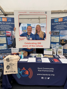Gary and Vicky gold a Railway 200 themed selfie frame at the Kent Community Rail Partnership promotional stall. Their are Kent CRP roller banners either side and an array of brochures and give-away items on the table.