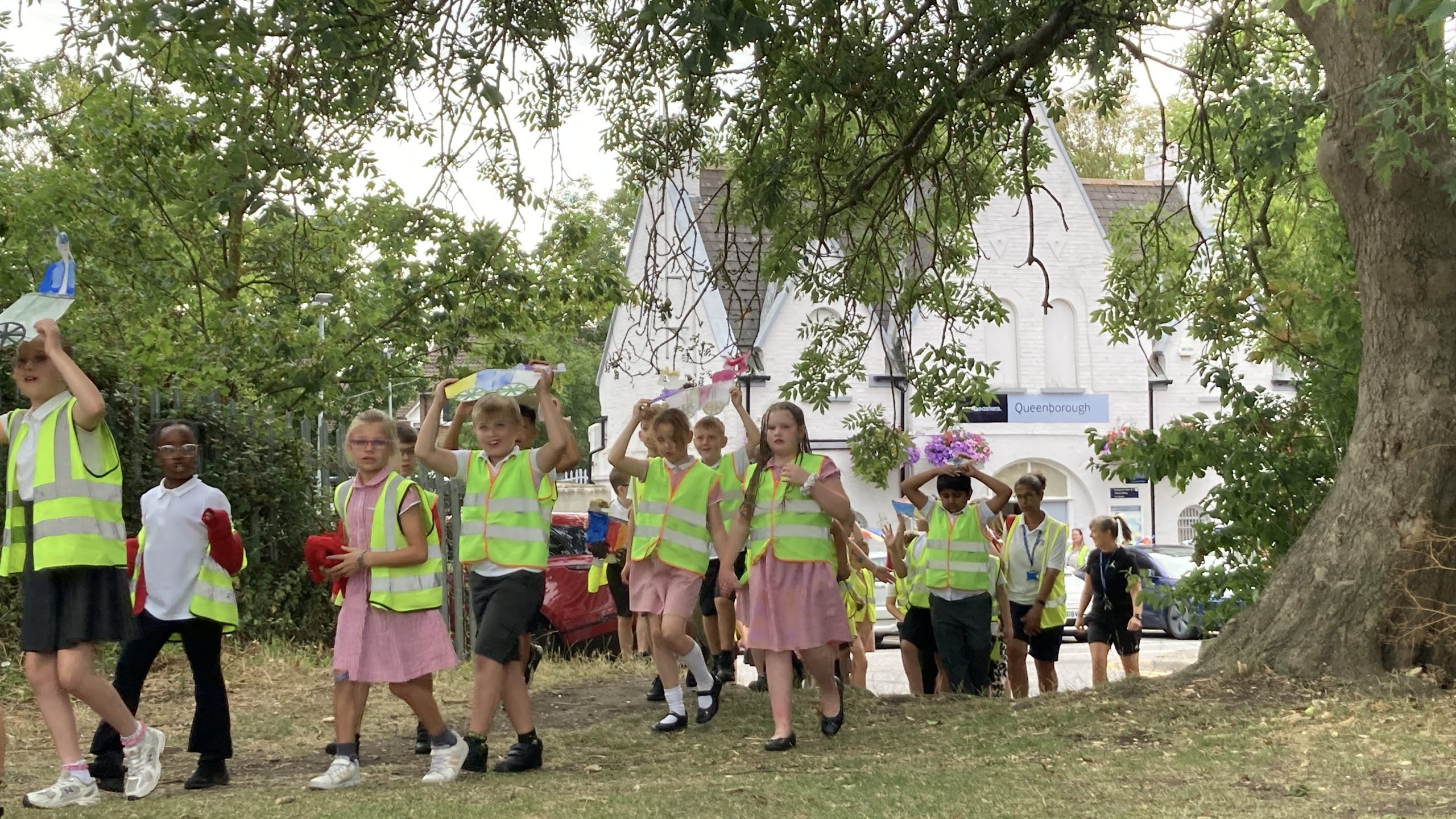 A group of children in hi-vis tops walking in procession onto the town green, holding colourful drawings of steam engines and carriages. Queenborough station building can be seen in the background
