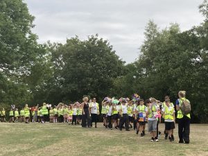 A group of children in hi-vis tops walking in procession across the town green, holding colourful drawings of steam engines and carriages