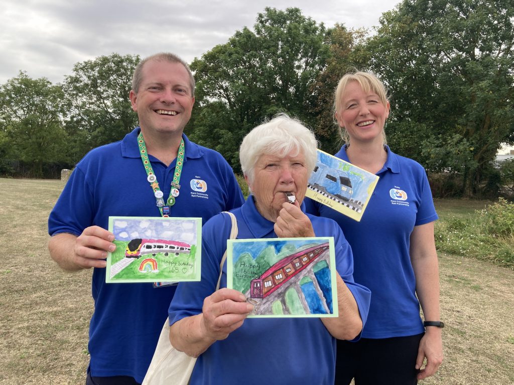 Gary, Linda and Vicky in blue Kent Community Rail Partnership top pose for a picture on the town green at Queenborough, holding birthday cards to celebrate the 165th birthday of Queenborough station. Linda is blowing a whistle