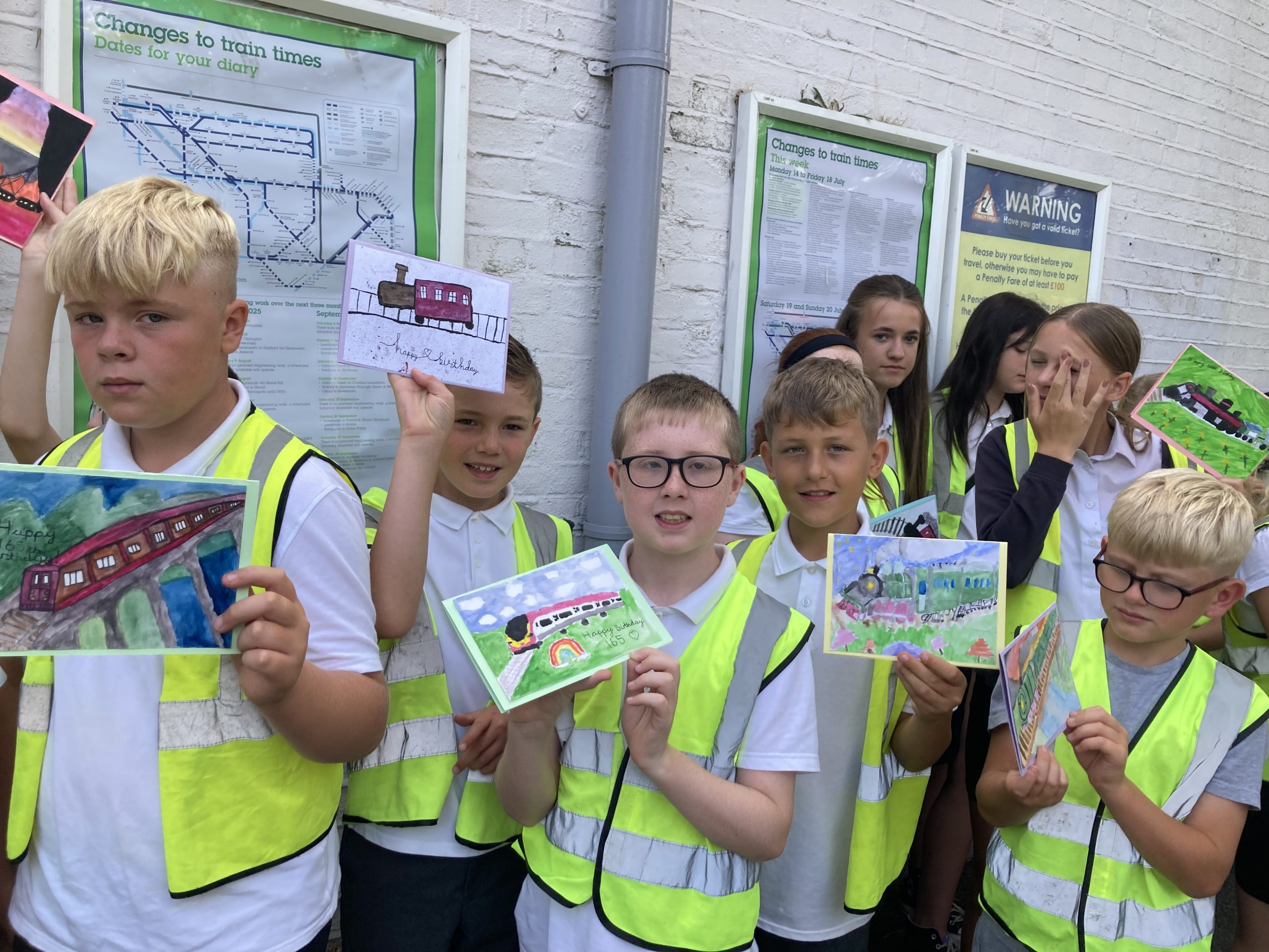 Young learners in hi-vis jackets display birthday cards they have created to celebrate Queenborough station's birthday