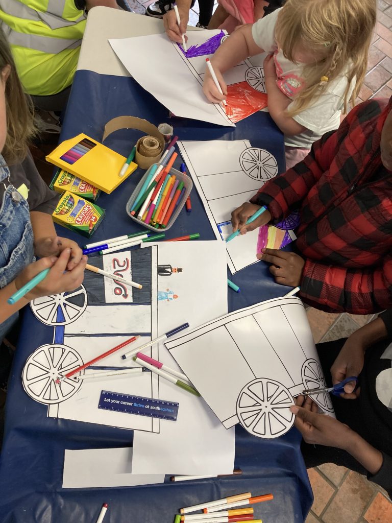 The hands of many children cutting our and colouring-in railway carriages on a table full of multi-coloured colouring-pens