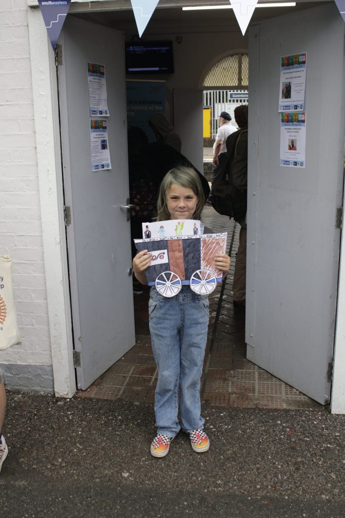 A young person holding their decorated train-carriage and passengers poses for a picture outside Queenborough station