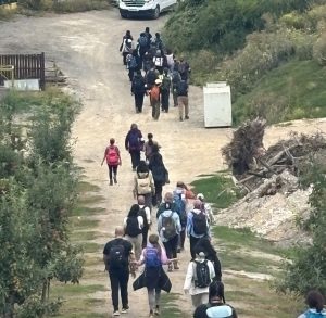 A large group of walkers, three or four abreast, head along a path away from camera. There is an orchard to one side, with rough ground, dead tress and a large white box to the other.