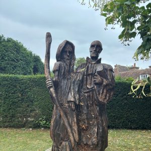 Two carved pilgrim figures on a neat green lawn in front of a trimmed hedge.