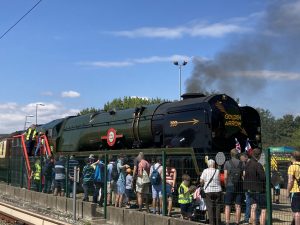 A line of people behind a fence, queuing to visit the driver's cab of Clan Line, a large green steam locomotive. Clan Line has a "Golden Arrow" train service logo on the front.