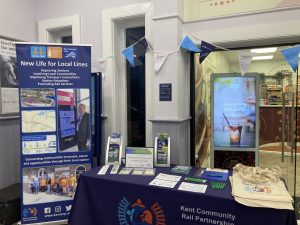 Kent Community Rail Partnership promotional stall in front of the station cafe. Colouring in sheets, pencils, tote bags and brochures are on the table. A roller banner is displayed to one side.