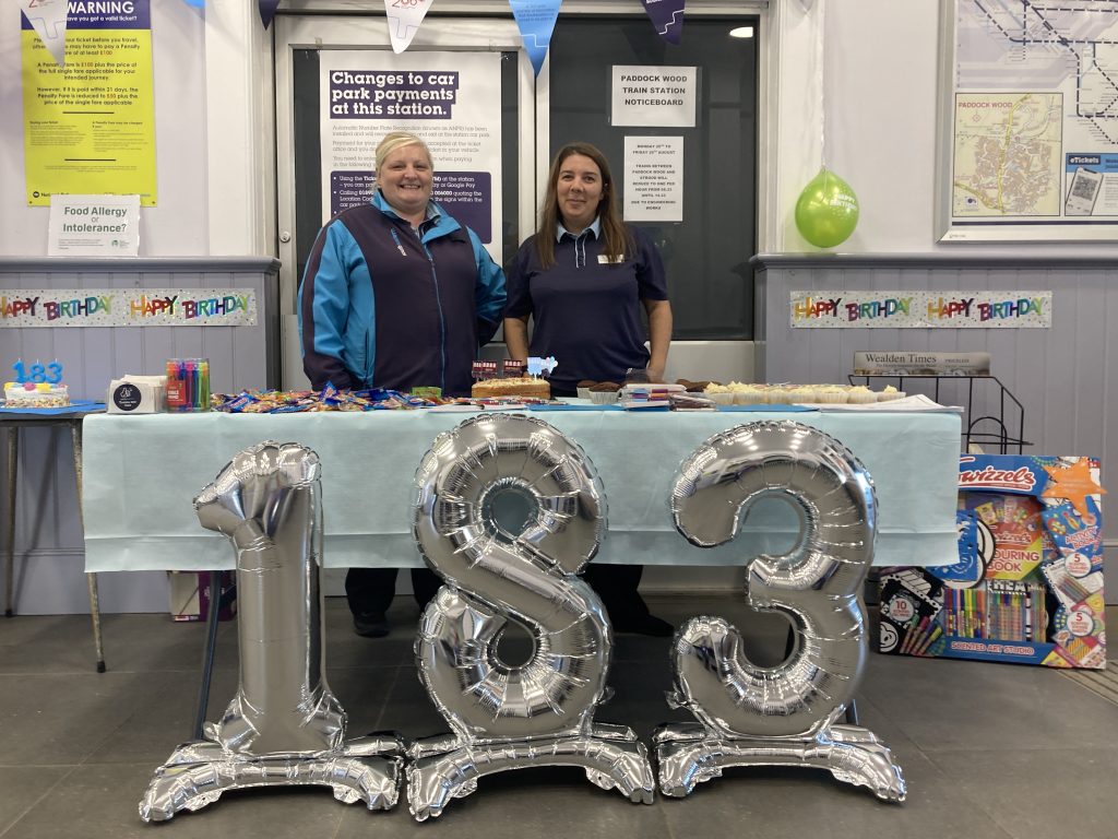 Clare and Choe stood behind a table loaded with sweets, cakes and giveaways. In front of the table are large silver balloon numbers, "183".