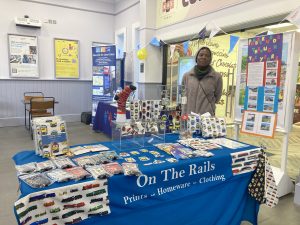 Neng Mbah stood behing the "On The Rails" table of rail related merchandise in the station booking hall. Beside the stall is an information panel "Paddock Wood through the years" displaying details of the station's history
