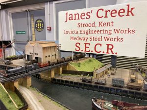 A diorama display of boats on the river beside wharves, industrial buildings, a railway line and sidings. "Janes' Creek, Strood, Kent. Invicta Engineering Wrls, Medway Steel works, South Eastern and Chatham Railway"