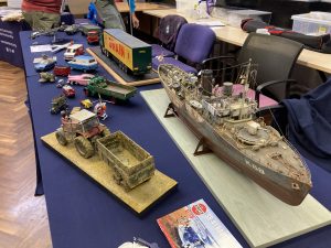 A variety of models from the Medway Modelling Club displayed on a table. A small naval ship, a tractor and trailer, various trucks vans and cars.