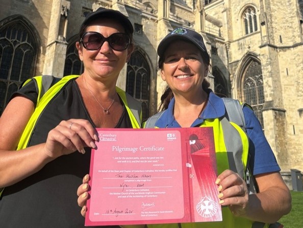 Two people hold a Pilgrimage certificate, they are standing in front of Canterbury cathedral