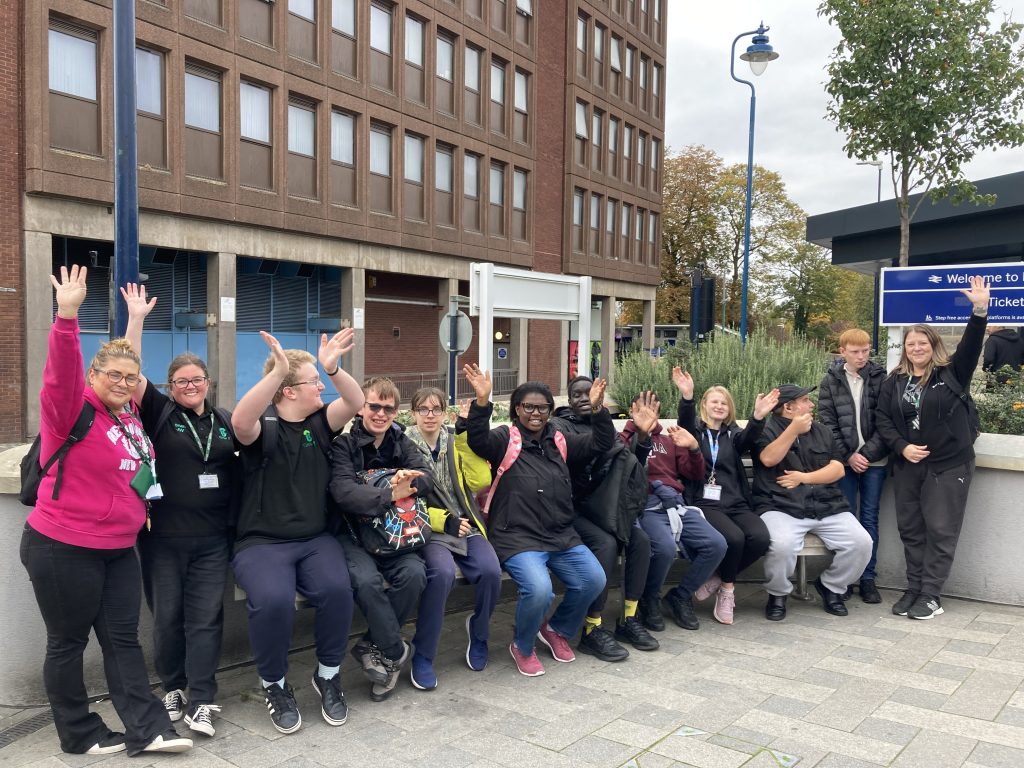 A group of people seated on a bench outside a station pose for a picture, waving at camera