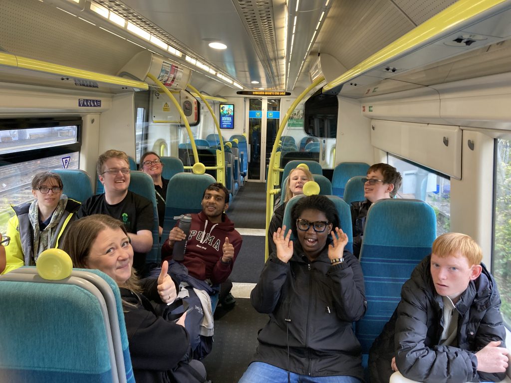A group of people seated on a train give a thumbs up to camera