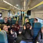 A group of people seated on a train give a thumbs up to camera