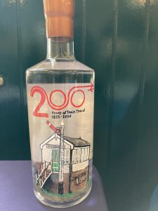 A bottle of clear liquid with an orange wrap around its neck. It is decorated with a celebratory Railway200 logo and a Signal Box