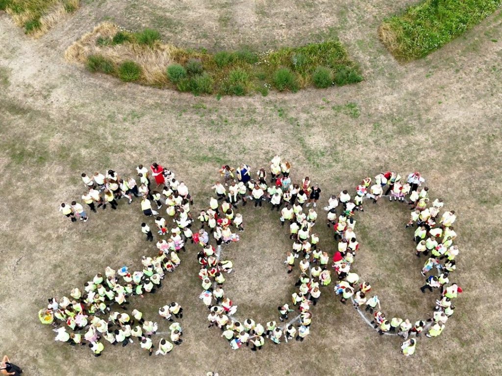 Drone image of over 200 schoolchildren in hi-vis tops on a village green. The children are assembled around chalk pain markings of the number two hundred.