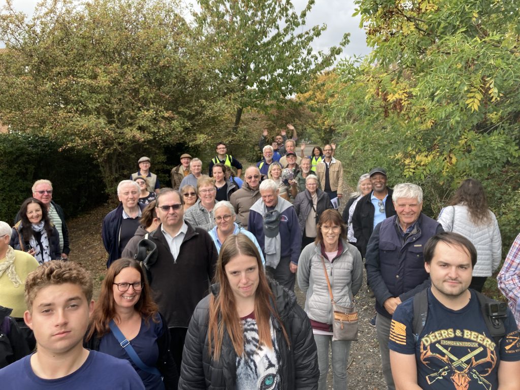 A crowd of people of a gravel path between trees pose for a photo. The people at the back are waving.