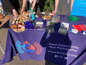 Kent Community Rail Partnership promotional stall with freebie brochures and pencils. Behind the stall students are offering delicious treats on silver foil trays