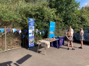 The Kent Community Rail partnership and Lenham school stands in the station carpark. The tables have tasty treats, brochures and freebie items. Behind the tables are banner displays and bunting. Two students and a tutor stand chatting nearby