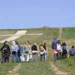 A group of walkers stand looking at a white chalk cross carved into a green hillside. Sky above is clear and blue.