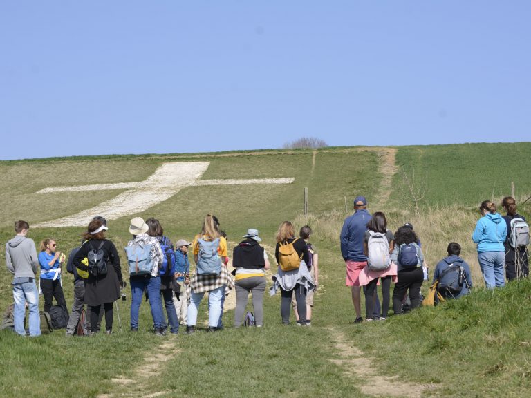 A group of walkers stand looking at a white chalk cross carved into a green hillside. Sky above is clear and blue.