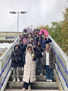 A crpwd of people pose for a photo on the steps of a station footbridge. It is raining, all are wearing coats, some have umbrellas.