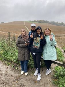 Four people pose for a photograph at the side of a gravel trail. Behind them is a ploughed field that climbs a hillside topped with trees