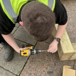 A person in hi-vis uses a power tool to screw wooden sleeper timbers together to construct a planter.