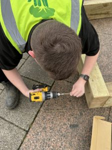 A person in hi-vis uses a power tool to screw wooden sleeper timbers together to construct a planter.