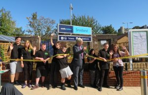 A group of people stand in a line holding a ribbon which one of the is cutting. They are waving in celebration. Behind them is the sign for Snodland station and a newly planted garden