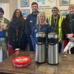 A group of sic people pose for a photo in Charing station booking office. In front of them on a table are two large urns of tea and coffee and a tub of chocolate sweets
