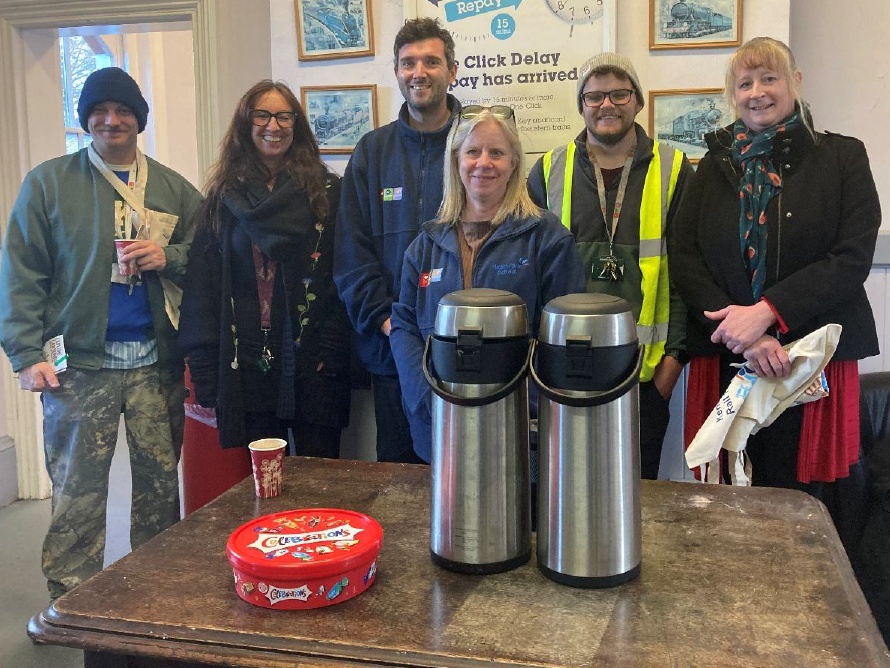 A group of sic people pose for a photo in Charing station booking office. In front of them on a table are two large urns of tea and coffee and a tub of chocolate sweets