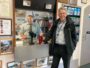 Mark behind the glass of the ticket office and Matt in front pose for a photo.