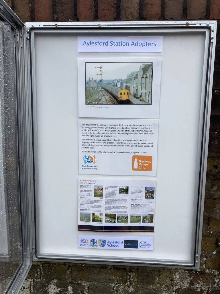 An open poster cabinet headed "Aylesford Station Adopters" displays a colour image of a slam-door train the station and details of its history. A poster gives information about places to visit on the Medway Valley Line, Logos of Kent Community Rail Partnership, Aylesford Parish Council, Aylesford Schoo;, Southeastern and Community Rail Network Funded.