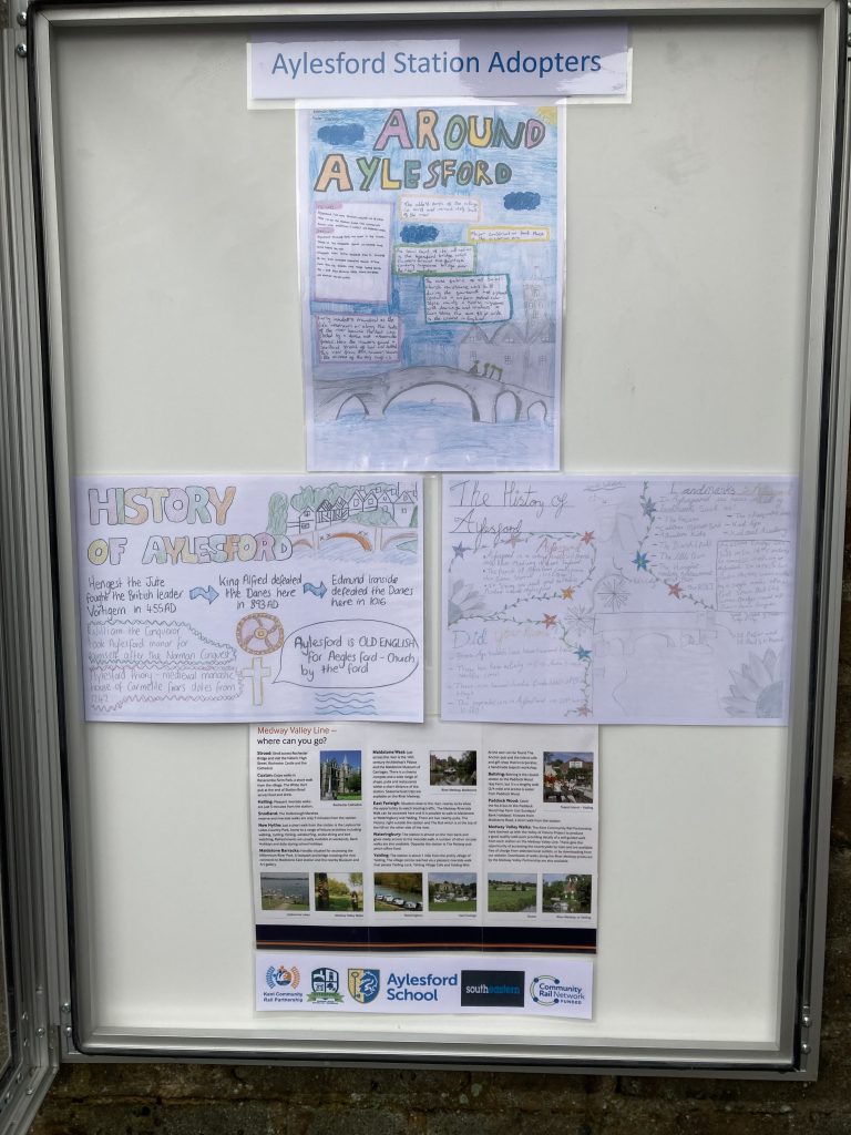 An open poster cabinet headed "Aylesford Station Adopters" displays student artwork about Aylesford village and its history. A poster gives information about places to visit on the Medway Valley Line, Logos of Kent Community Rail Partnership, Aylesford Parish Council, Aylesford Schoo;, Southeastern and Community Rail Network Funded.