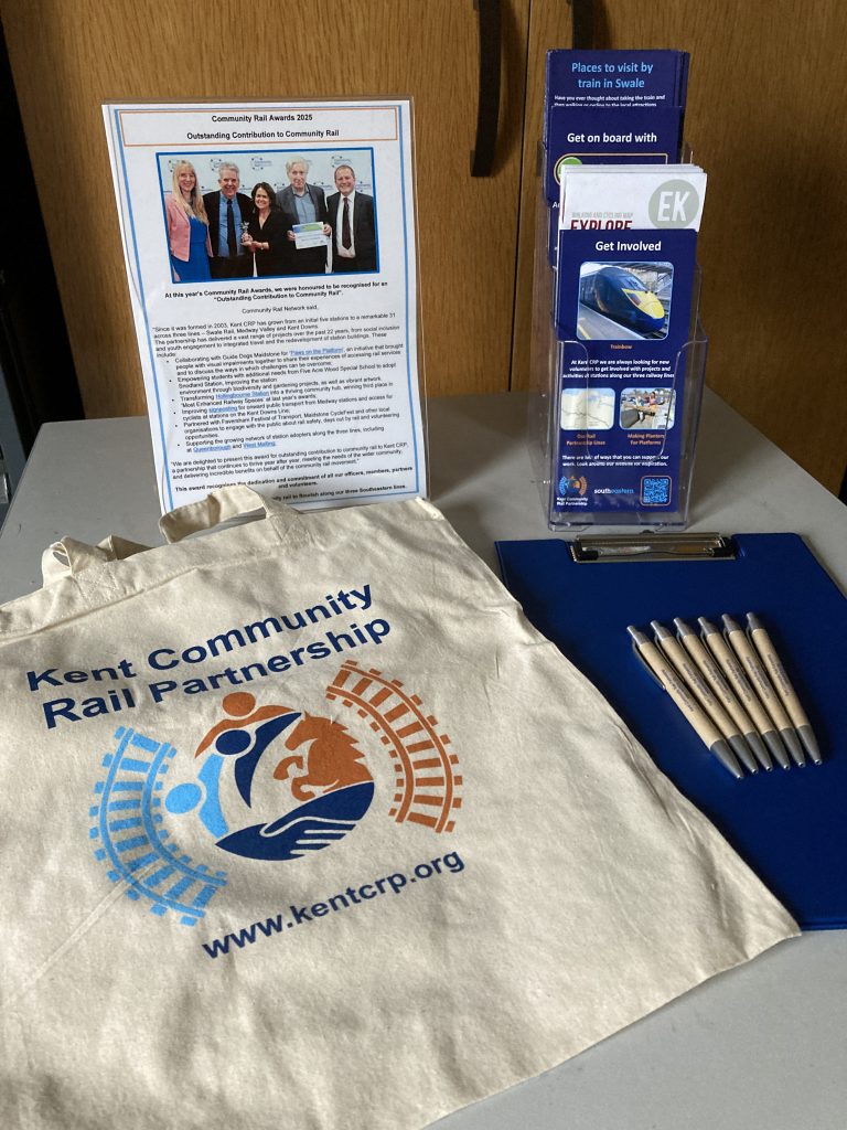 An A4 poster display with a notice about Kent Community Rail Partnership's "Outstanding Contribution to Community Rail" award for 2025, A leaflet dispenser full of brochures, a Kent Community Rail Partnership branded tote bag, clipboard and Kent CRP branded pens on a table.