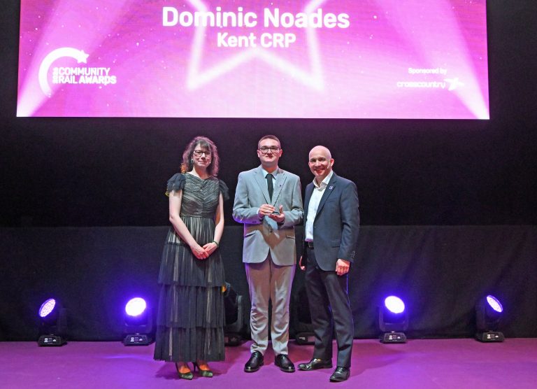 Dominic Noades on stage at the Community Rail Awards 2026. He is receiving his award from the hosts of the evening.