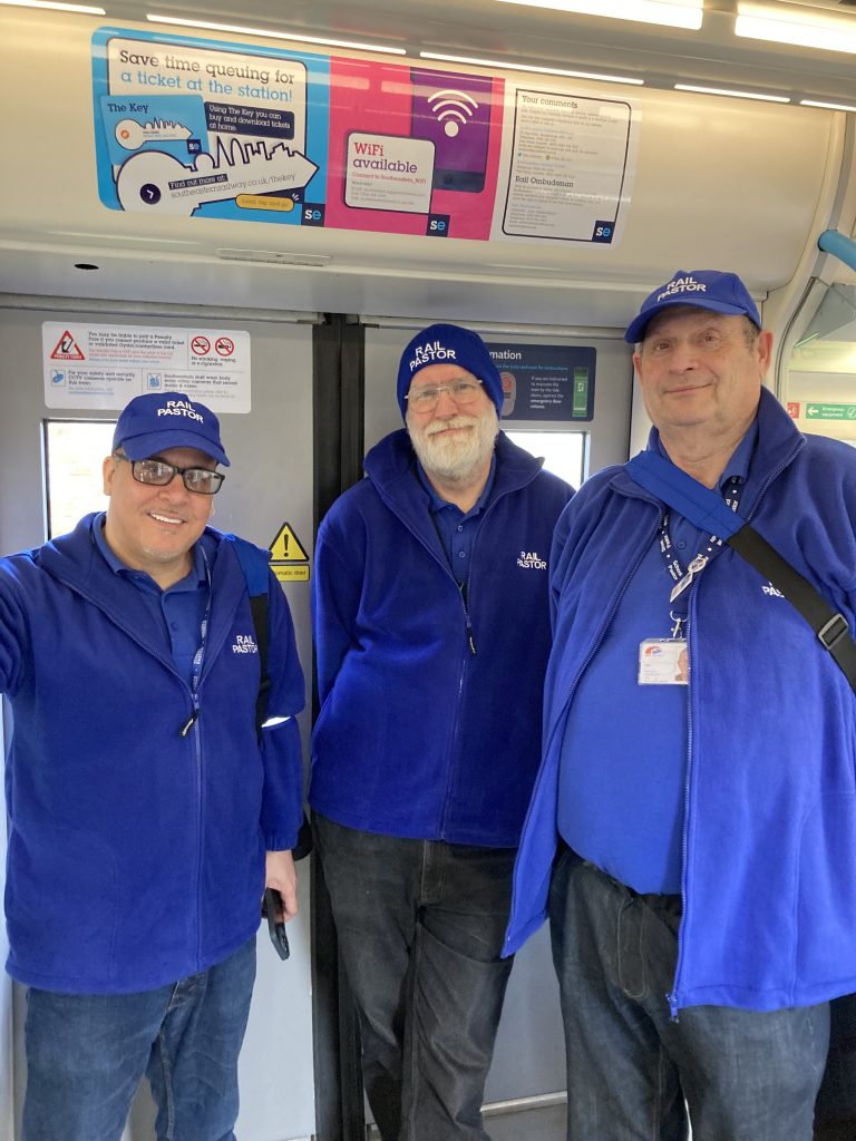 Three people in matching blue hats, jackets and tops with a "Rail Pastor" logo pose for a picture in the door vestibule onboard a train.
